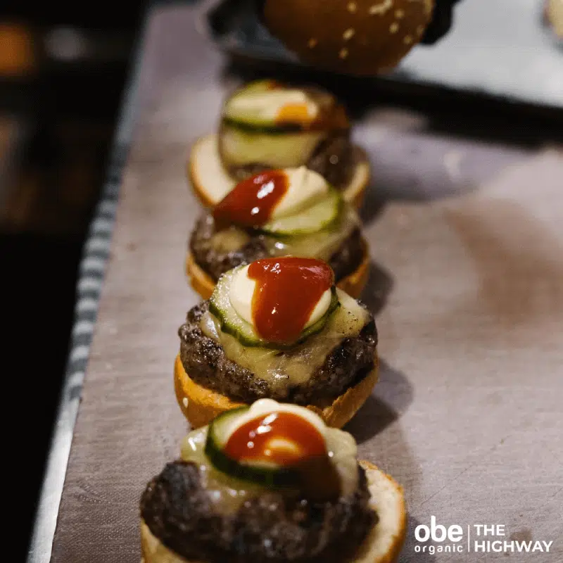 Row of mini burgers with colorful toppings on a stainless tray.