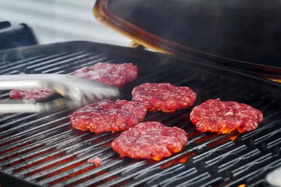 Raw beef burger patties sizzling on a hot grill with metal tongs flipping one patty.