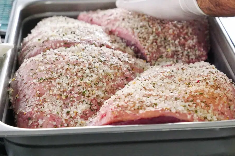 Several cuts of raw beef coated with coarse salt and herbs in a metal tray, with a gloved hand preparing the meat.