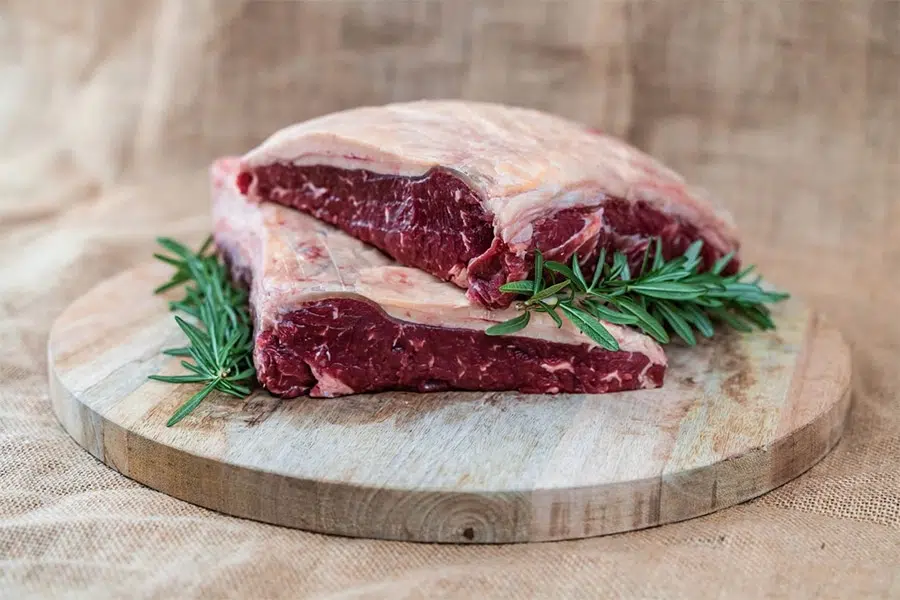 Two raw beef cuts with a fat cap, garnished with fresh rosemary, on a round wooden cutting board.