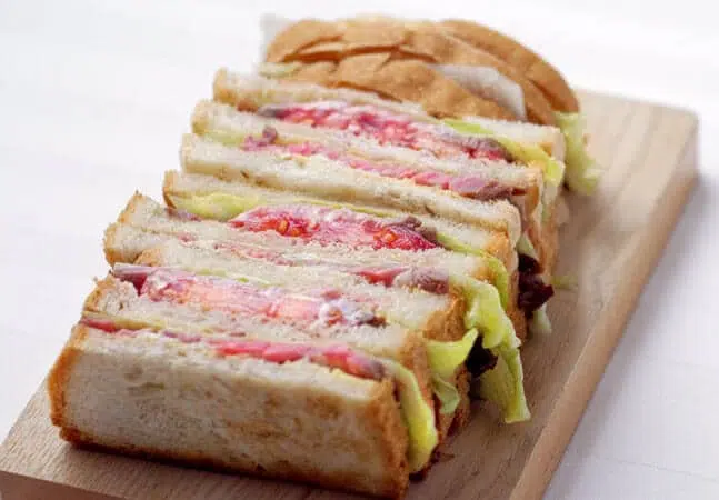 Sliced sandwich loaf with layers of lettuce, tomato, and beef between thick slices of bread, arranged on a wooden board.
