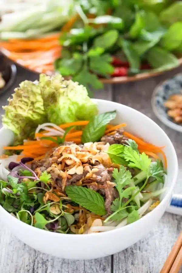 Bun Bo Xao Goi Bo Vien Viet Nam Vietnamese noodle bowl with grilled beef, fresh herbs, shredded carrots, lettuce, and crispy shallots served in a white bowl.