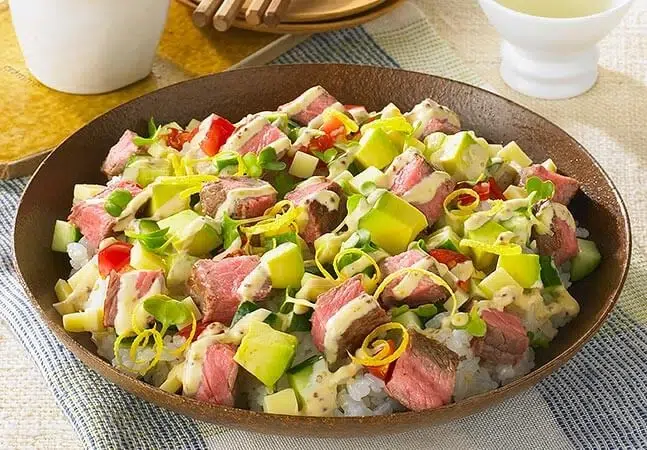 Bowl of sushi rice topped with diced seared beef, avocado, red peppers, green onions, lemon zest, and drizzled with a creamy sauce.