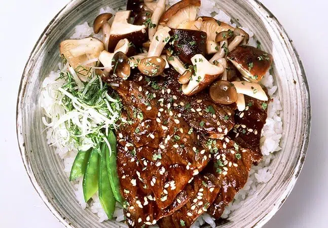 Bowl of rice topped with glazed beef slices, assorted mushrooms, snap peas, shredded green onions, and sesame seeds.