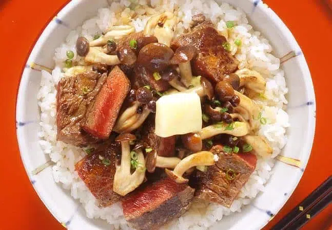 Bowl of rice topped with seared beef cubes, sautéed mushrooms, chopped green onions, and a pat of butter.