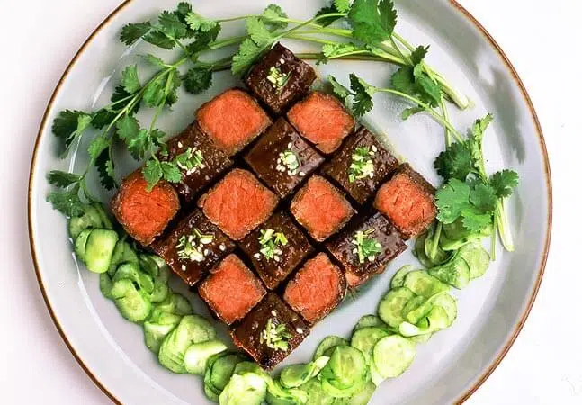 Plate of neatly arranged beef cubes topped with green garnish, surrounded by cucumber slices and fresh cilantro.