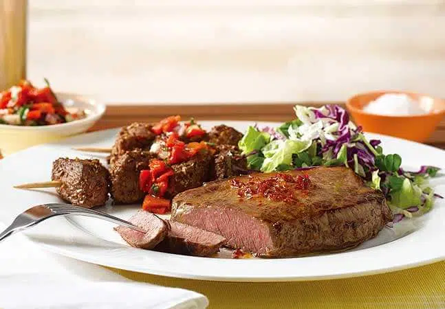 Plate with seared steak, beef skewers topped with red pepper salsa, and a side of fresh cabbage salad.