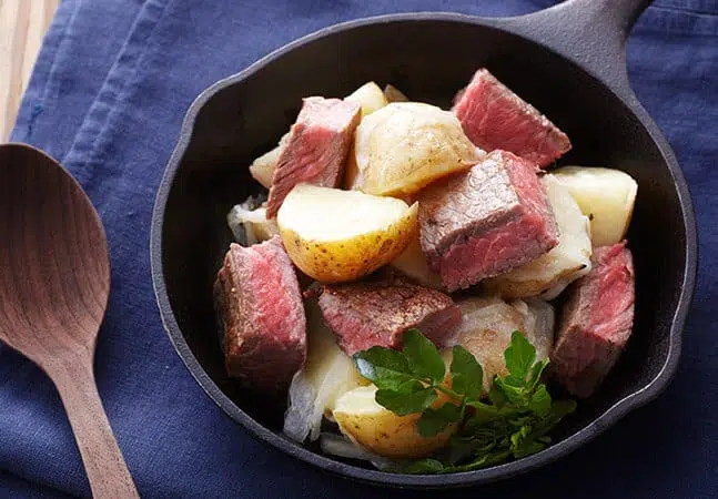 Skillet dish with seared beef cubes, roasted potatoes, onions, and a sprig of greens.
