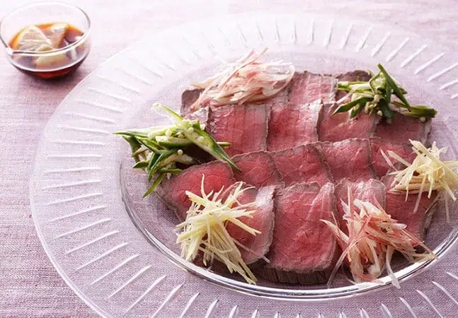 Thin slices of roast beef served on a glass plate with garnishes of ginger, green beans, and myoga, accompanied by dipping sauce.