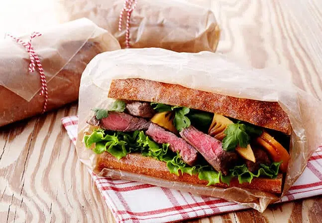 Steak sandwich with lettuce, tomato, herbs, and cheese wrapped in parchment paper on a wooden surface.
