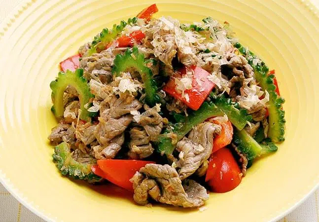 Plate of stir-fried beef with bitter melon, red bell peppers, and bonito flakes on top.