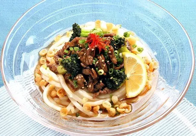 Bowl of udon noodles topped with beef, broccoli, green onions, sesame seeds, crispy tempura bits, and a lemon wedge.