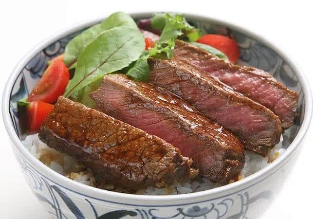 Bowl of rice topped with seared beef slices, fresh greens, and cherry tomato halves