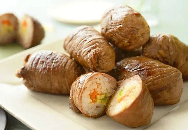 Plate of beef-wrapped rice rolls filled with vegetables, with one cut open to show rice, carrot, and green filling inside.