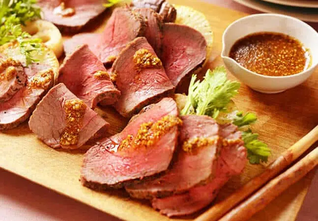 Sliced roast beef served on a wooden board with mustard sauce and garnished with greens.