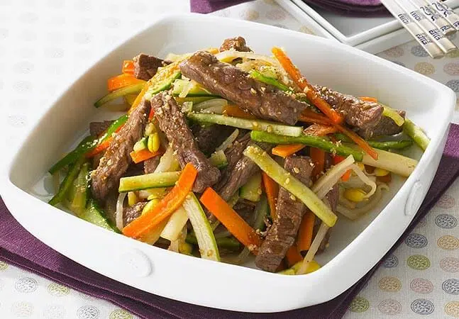 Stir-fried beef strips with carrots, cucumbers, bean sprouts, and sesame seeds served in a white dish.