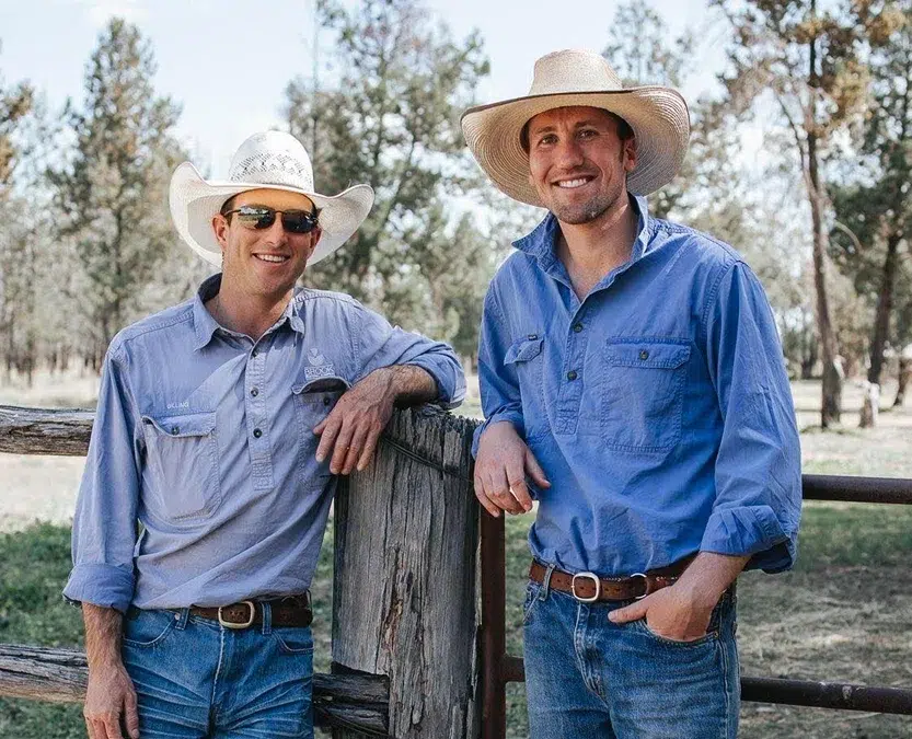 Two smiling men in cowboy hats and casual work shirts stand outdoors by a wooden fence on a ranch, surrounded by trees.
